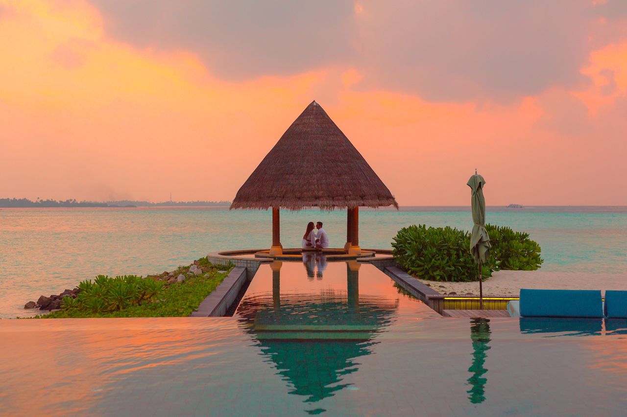 menu-01 A couple enjoying a romantic moment by the sea and pool under a beach cabana during sunset.
