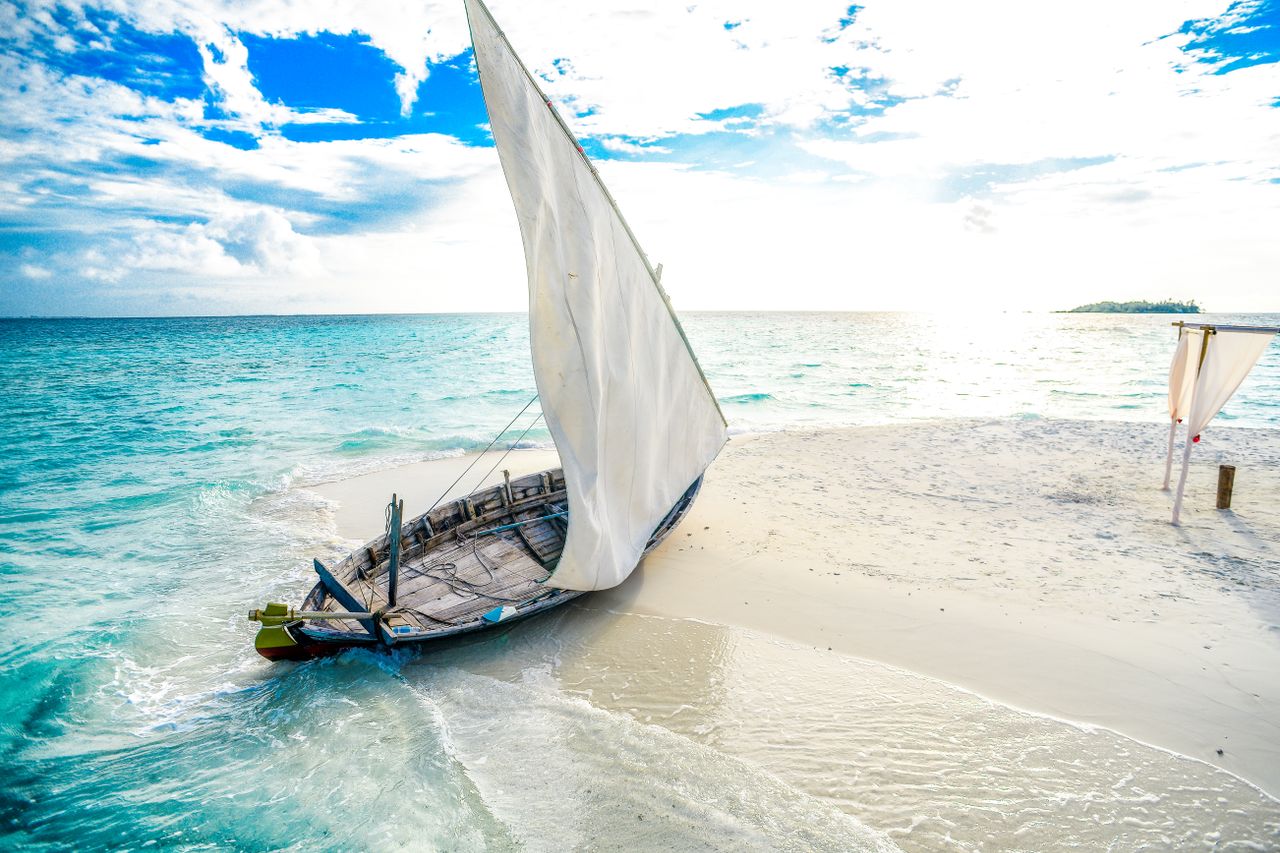 Menu A picturesque sailboat rests on the white sand of a tropical island beach with turquoise waters and clear skies.
